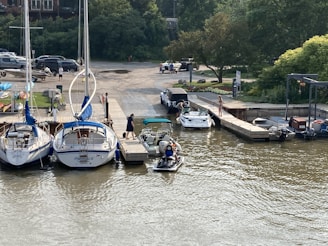 A marina with several boats docked in the water. People can be seen near the boats, with one person standing on the dock, and others preparing a small boat. Trees and parked cars are visible in the background.