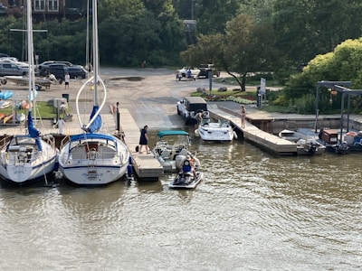 A marina with several boats docked in the water. People can be seen near the boats, with one person standing on the dock, and others preparing a small boat. Trees and parked cars are visible in the background.