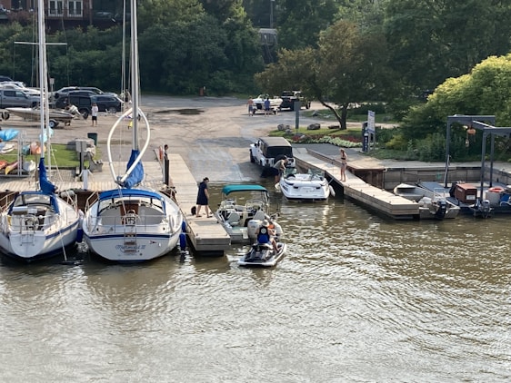 A marina with several boats docked in the water. People can be seen near the boats, with one person standing on the dock, and others preparing a small boat. Trees and parked cars are visible in the background.