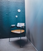 A modern chair with a minimalist design stands in a corner with a textured blue-green wall. The chair features a wooden seat and backrest with teal metal legs and armrests. Two round white wall decorations are aligned vertically on the wall.