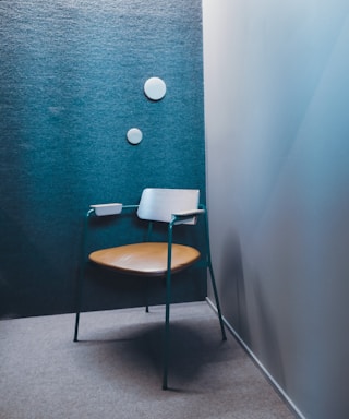 A modern chair with a minimalist design stands in a corner with a textured blue-green wall. The chair features a wooden seat and backrest with teal metal legs and armrests. Two round white wall decorations are aligned vertically on the wall.