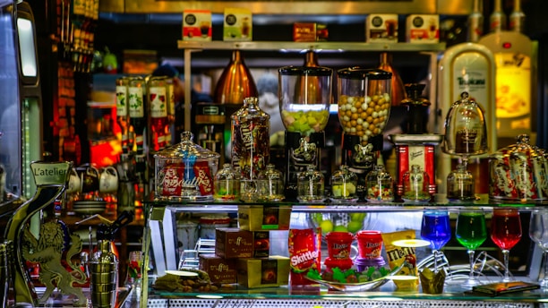 A vibrant display of assorted candies and snacks is arranged on a shelf. Various glass jars filled with treats like colorful candies and chocolates are visible, alongside brand packaging for products such as Skittles and M&Ms. Illuminated by colorful lighting reflecting on glass containers, the setup conveys a sense of indulgence and variety.