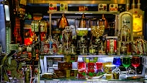 A vibrant display of assorted candies and snacks is arranged on a shelf. Various glass jars filled with treats like colorful candies and chocolates are visible, alongside brand packaging for products such as Skittles and M&Ms. Illuminated by colorful lighting reflecting on glass containers, the setup conveys a sense of indulgence and variety.