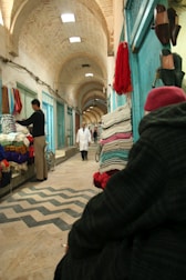 An indoor marketplace with arched ceilings, vendors displaying colorful textiles, and a person in traditional clothing walking down the aisle. Brightly colored fabrics are stacked against teal walls, and shoes hang on the right side. A man stands by his textiles on the left, while a cloaked figure with a red hat sits in the foreground.