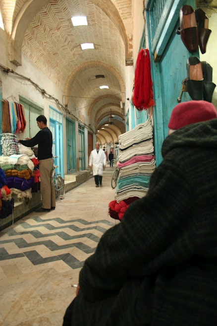 An indoor marketplace with arched ceilings, vendors displaying colorful textiles, and a person in traditional clothing walking down the aisle. Brightly colored fabrics are stacked against teal walls, and shoes hang on the right side. A man stands by his textiles on the left, while a cloaked figure with a red hat sits in the foreground.