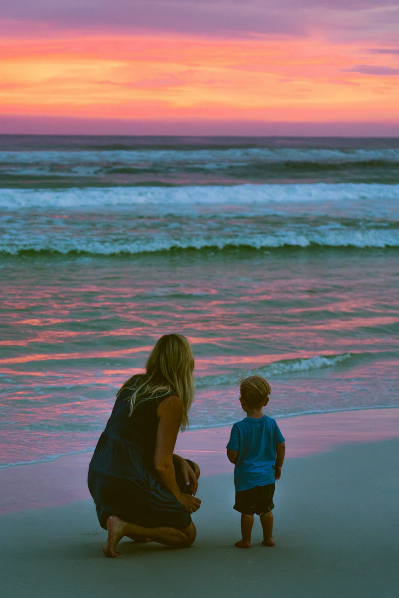 Mãe agachada ao lado do filho diante do mar ao pôr do sol rosado