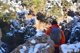 A person wearing a bright orange jacket and a backpack is seen from behind, navigating a snow-dusted rocky terrain, surrounded by trees partially covered in snow. The scene suggests a winter hiking or trekking expedition through a rugged landscape.
