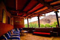 A rustic outdoor seating area with wooden beams supporting a thatched roof, featuring a mix of sofas and tables. The interior includes patterned cushions and a combination of clay and natural wood textures. The view extends to a mountainous landscape with lush greenery in the foreground.