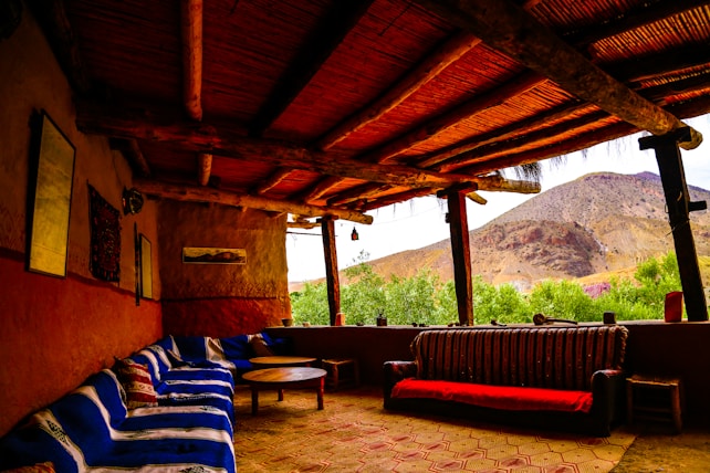 A rustic outdoor seating area with wooden beams supporting a thatched roof, featuring a mix of sofas and tables. The interior includes patterned cushions and a combination of clay and natural wood textures. The view extends to a mountainous landscape with lush greenery in the foreground.