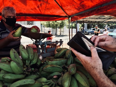 A vendor wearing a mask is holding and weighing green plantains at a market stall. Another person is holding an open wallet, preparing to pay. The stall is covered by a red canopy, and there are more customers in the background. The scene appears to be in an outdoor market setting.