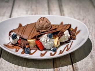 A delicious chocolate dessert crepe filled with whipped cream, bananas, and strawberries, topped with a scoop of chocolate ice cream and drizzled with chocolate syrup. Blueberries and crumbled brownie pieces add texture and sweetness.