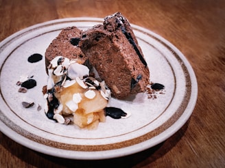 Two slices of rich chocolate dessert are elegantly arranged on a plate. Beside them, there's a dollop of whipped cream topped with sliced almonds and drizzled with chocolate sauce. The dessert is placed on a textured, ceramic plate with a subtle decorative rim, and the setting is on a wooden table.