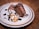Two slices of rich chocolate dessert are elegantly arranged on a plate. Beside them, there's a dollop of whipped cream topped with sliced almonds and drizzled with chocolate sauce. The dessert is placed on a textured, ceramic plate with a subtle decorative rim, and the setting is on a wooden table.