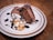 Two slices of rich chocolate dessert are elegantly arranged on a plate. Beside them, there's a dollop of whipped cream topped with sliced almonds and drizzled with chocolate sauce. The dessert is placed on a textured, ceramic plate with a subtle decorative rim, and the setting is on a wooden table.