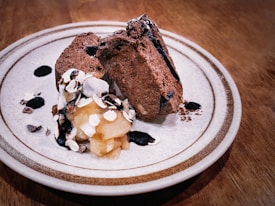 Two slices of rich chocolate dessert are elegantly arranged on a plate. Beside them, there's a dollop of whipped cream topped with sliced almonds and drizzled with chocolate sauce. The dessert is placed on a textured, ceramic plate with a subtle decorative rim, and the setting is on a wooden table.