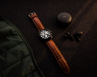 A brown leather-strapped wristwatch and a set of black wireless earbuds with their charging case are placed on a dark fabric surface. There is a green quilted jacket partially visible on the left side.