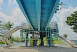 A vibrant playground structure featuring metal slides and a climbable grid surface is situated under a large blue overhead walkway. The surrounding area is sandy with patches of grass, and lush green trees are visible against a bright, partly cloudy sky.