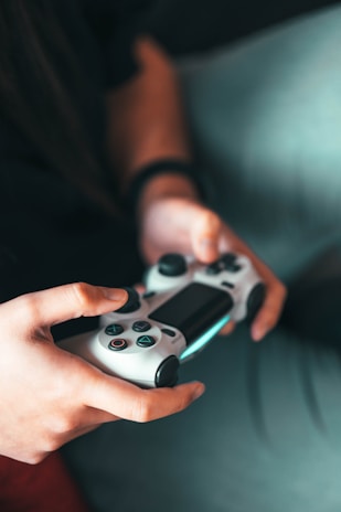 A person is holding a white video game controller with their thumbs on the buttons, in a comfortable indoor setting. The light is soft, creating a warm and focused atmosphere around the hands and the controller.