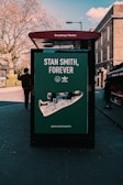 A street advertisement for Adidas featuring a large image of a Stan Smith shoe displayed on a bus stop shelter. The advertisement is located at Broadway Market. There is a person walking past, and the background includes bare trees and some brick buildings.