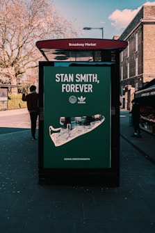 A street advertisement for Adidas featuring a large image of a Stan Smith shoe displayed on a bus stop shelter. The advertisement is located at Broadway Market. There is a person walking past, and the background includes bare trees and some brick buildings.