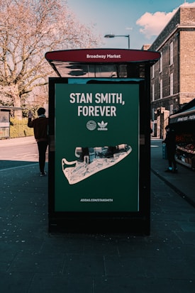 A street advertisement for Adidas featuring a large image of a Stan Smith shoe displayed on a bus stop shelter. The advertisement is located at Broadway Market. There is a person walking past, and the background includes bare trees and some brick buildings.