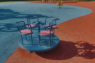 A playground area with a round blue and red merry-go-round featuring four seats with handlebars. The ground is covered in blue and red rubberized surface, with a small yellow rocking toy visible in the background.