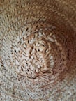 A close-up view of a woven straw hat with intricate patterns and a natural texture. The focus is on the center of the hat, highlighting the craftsmanship and details of the weave.