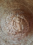 A close-up view of a woven straw hat with intricate patterns and a natural texture. The focus is on the center of the hat, highlighting the craftsmanship and details of the weave.