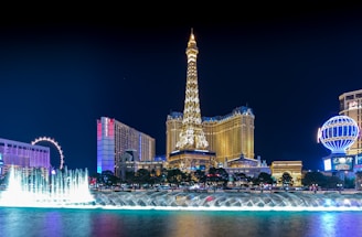 Illuminated nighttime cityscape featuring a replica of the Eiffel Tower prominently centered, surrounded by a skyline of glitzy hotels and casinos. A vibrant fountain display is in the foreground, with water jets creatively lit up.