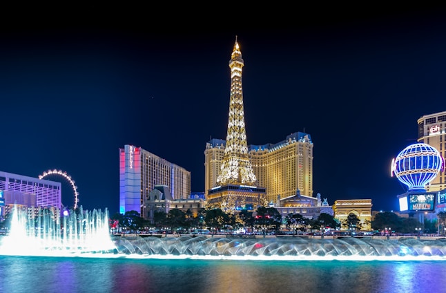 Illuminated nighttime cityscape featuring a replica of the Eiffel Tower prominently centered, surrounded by a skyline of glitzy hotels and casinos. A vibrant fountain display is in the foreground, with water jets creatively lit up.