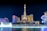 Illuminated nighttime cityscape featuring a replica of the Eiffel Tower prominently centered, surrounded by a skyline of glitzy hotels and casinos. A vibrant fountain display is in the foreground, with water jets creatively lit up.