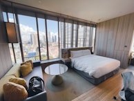 A modern hotel room features a large bed with white linens, a corner sofa with cushions, and a round coffee table. A large wall of windows provides a panoramic view of the city skyline with various skyscrapers.