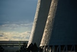 Tall, industrial cooling towers rise against a backdrop of cloudy skies. The sunlight casts patterns of light and shadow on the towers, giving them a textured appearance. In the foreground, silhouettes of trees and a building are visible.
