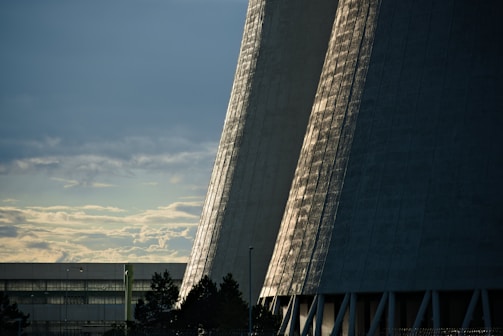 Tall, industrial cooling towers rise against a backdrop of cloudy skies. The sunlight casts patterns of light and shadow on the towers, giving them a textured appearance. In the foreground, silhouettes of trees and a building are visible.