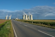 A rural road leads towards a power plant with large cooling towers emitting steam into a partially cloudy sky. The surrounding area consists of farmland with green fields and sparse vegetation lining the road.
