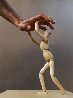 A wooden mannequin is positioned as if it's being supported or helped up by a larger human hand. The hand appears to gently hold the mannequin's upper body, illustrating an act of assistance or guidance. The background is neutral and plain, emphasizing the interaction between the hand and the mannequin.