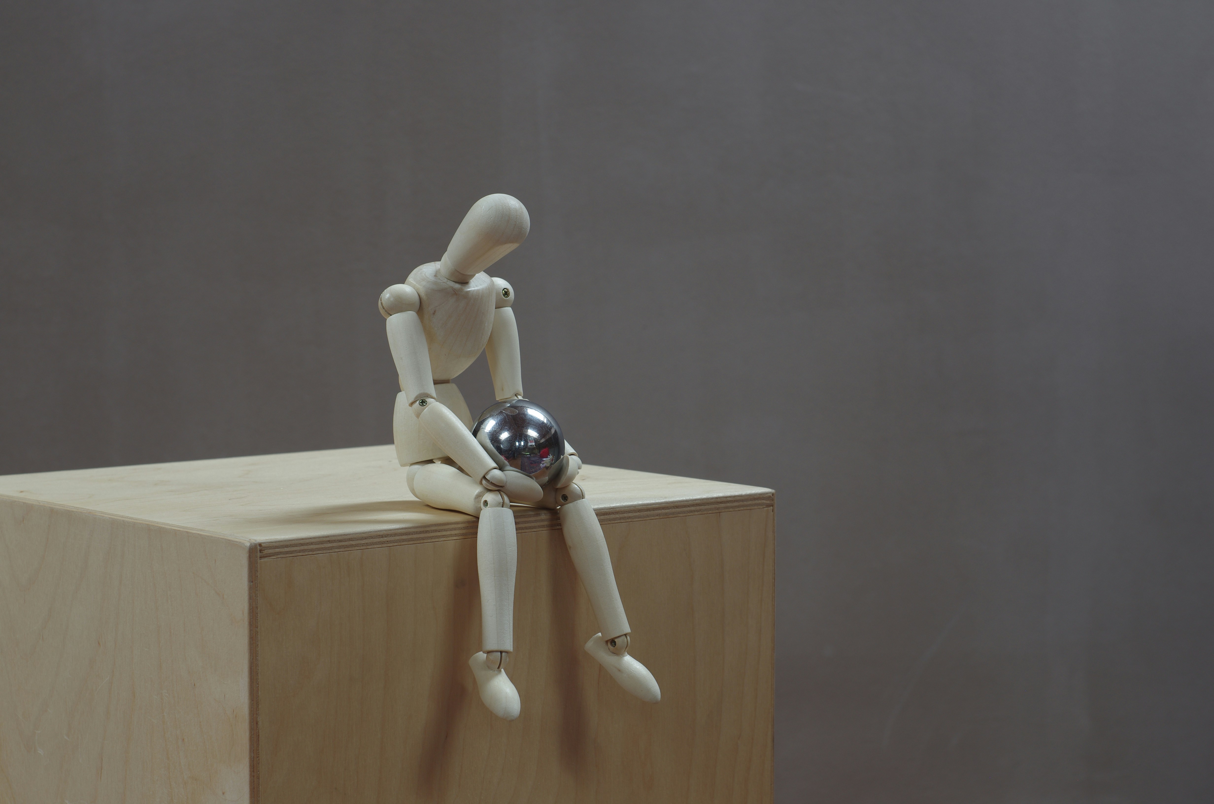 Seated wooden mannequin pensively observing a steel ball.