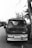 An old bus is parked on the side of the road near some trees. The bus is slightly worn, and the scene is depicted in black and white, giving it a nostalgic or vintage feel. The license plate is visible, and there are shadows of trees on the vehicle.