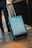 A person is seen walking, pulling a modern, hard-shell suitcase with wheels along a textured, metallic surface. The suitcase is sleek and compact, suggesting it is suitable for travel. The person's attire is casual, including dark pants and sneakers.