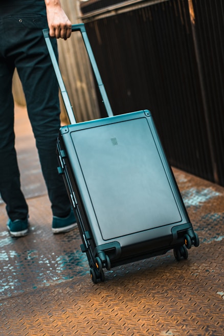 A person is seen walking, pulling a modern, hard-shell suitcase with wheels along a textured, metallic surface. The suitcase is sleek and compact, suggesting it is suitable for travel. The person's attire is casual, including dark pants and sneakers.