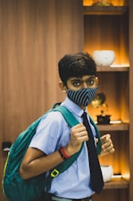 A young person wearing a blue uniform with a dark tie and a large green backpack. Their face is partially covered with a blue-striped mask. In the background, there are wooden shelves with decorative items, including vases, illuminated by warm lighting.
