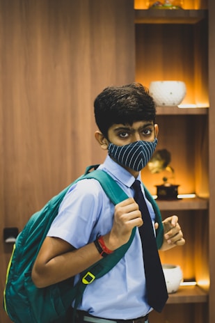 A young person wearing a blue uniform with a dark tie and a large green backpack. Their face is partially covered with a blue-striped mask. In the background, there are wooden shelves with decorative items, including vases, illuminated by warm lighting.