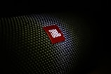 A close-up of a textured surface with a red, triangular logo displaying the letters 'JBL' in white. The texture resembles a woven pattern, commonly seen on speaker grills.