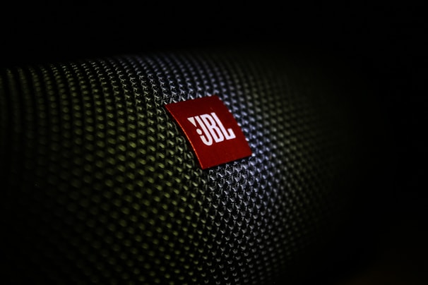 A close-up of a textured surface with a red, triangular logo displaying the letters 'JBL' in white. The texture resembles a woven pattern, commonly seen on speaker grills.