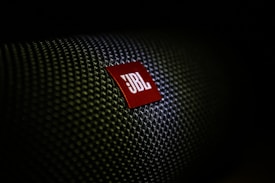 A close-up of a textured surface with a red, triangular logo displaying the letters 'JBL' in white. The texture resembles a woven pattern, commonly seen on speaker grills.
