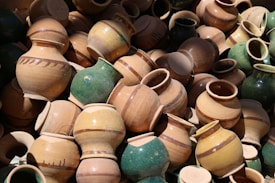 A large collection of traditional clay pots with varying earth tones and some with a green glaze. The pots are simple in design, with some featuring subtle decorative bands, and they are stacked in a casual, chaotic manner.