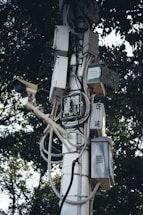 A tall pole is covered with multiple electrical boxes and surveillance cameras, intertwined with various wires and cables. The equipment is mounted in an organized manner, with trees in the background providing a natural contrast.