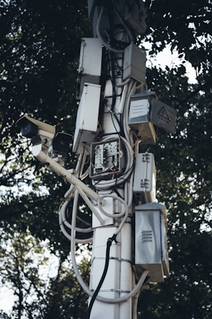 A tall pole is covered with multiple electrical boxes and surveillance cameras, intertwined with various wires and cables. The equipment is mounted in an organized manner, with trees in the background providing a natural contrast.