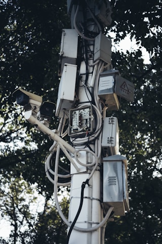 A tall pole is covered with multiple electrical boxes and surveillance cameras, intertwined with various wires and cables. The equipment is mounted in an organized manner, with trees in the background providing a natural contrast.