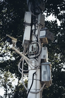 A tall pole is covered with multiple electrical boxes and surveillance cameras, intertwined with various wires and cables. The equipment is mounted in an organized manner, with trees in the background providing a natural contrast.